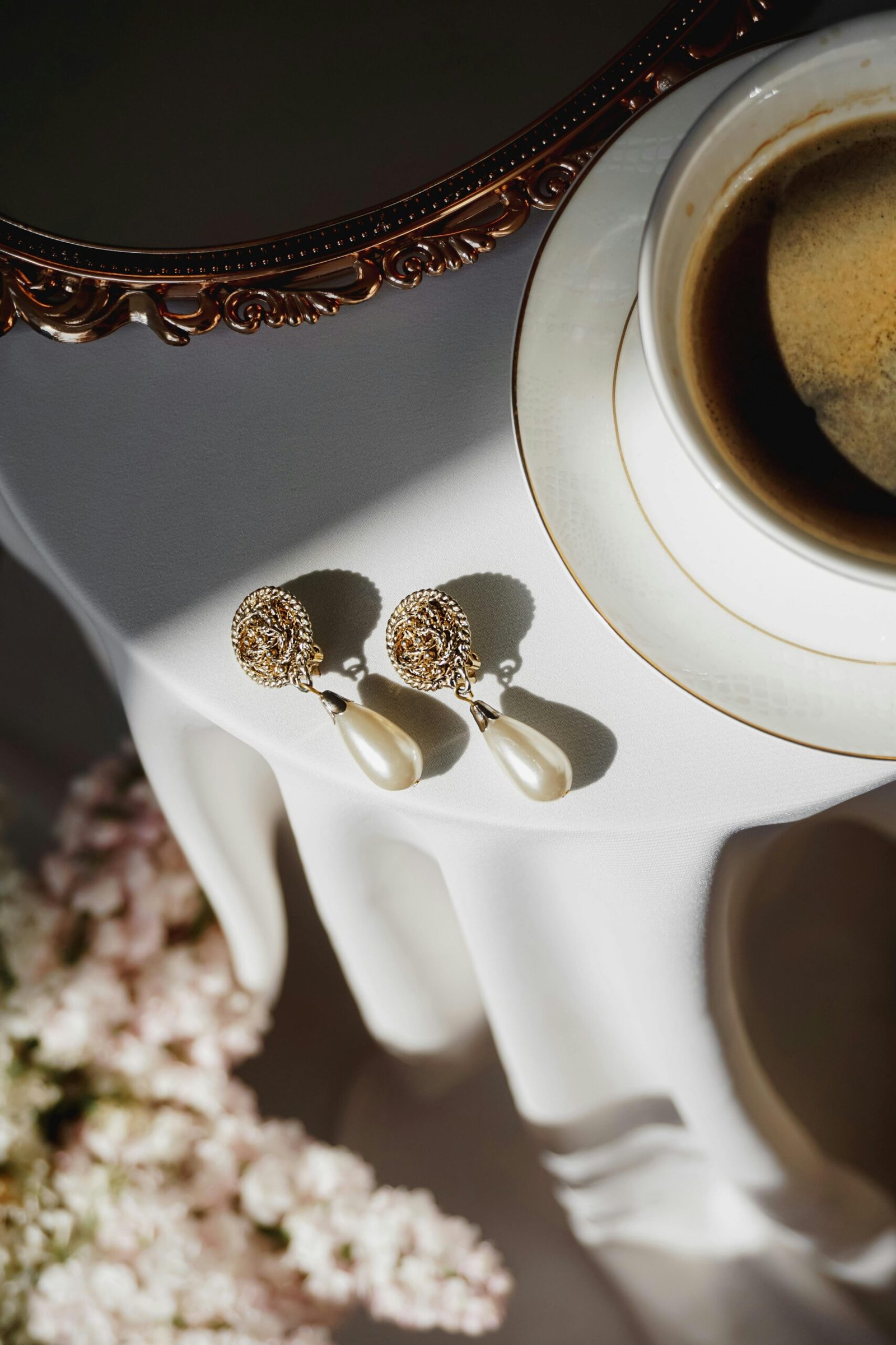 A pair of pearl earrings beside a cup of black coffee on a sunlit, vintage-style table.