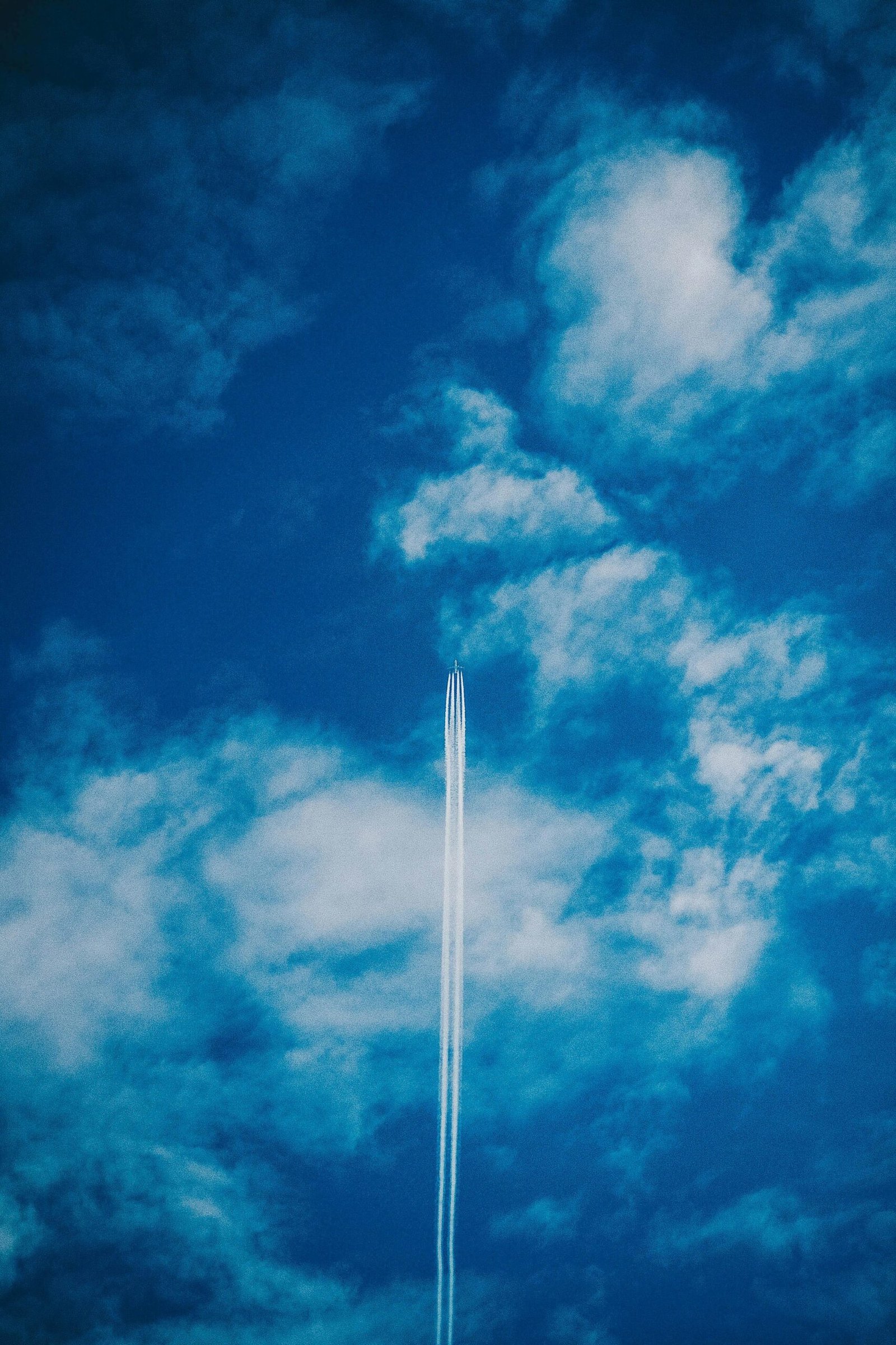 An airplane leaves a contrail across a bright blue sky filled with light clouds.