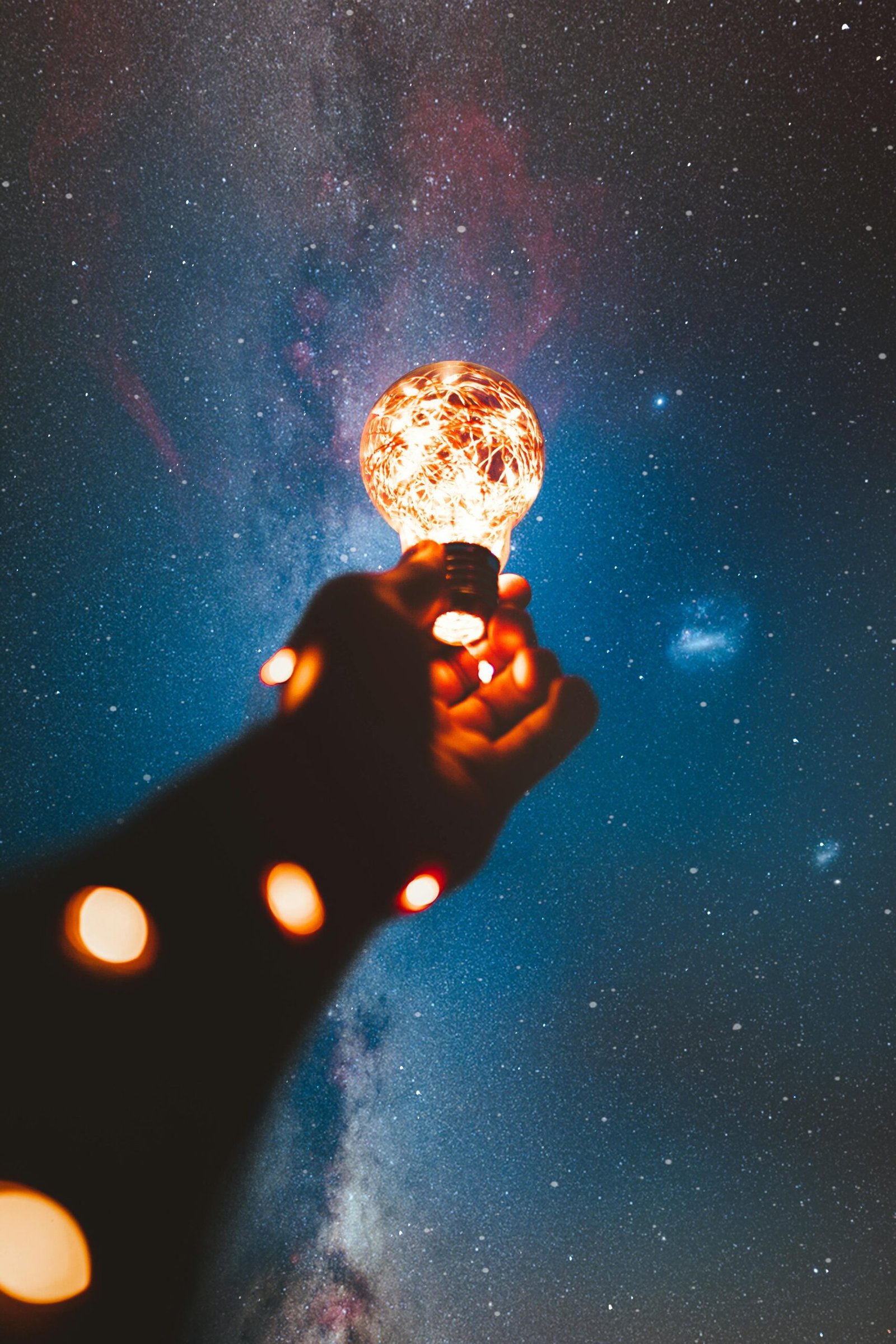 A hand holds a glowing bulb into the Milky Way night sky, creating a magical cosmic connection.