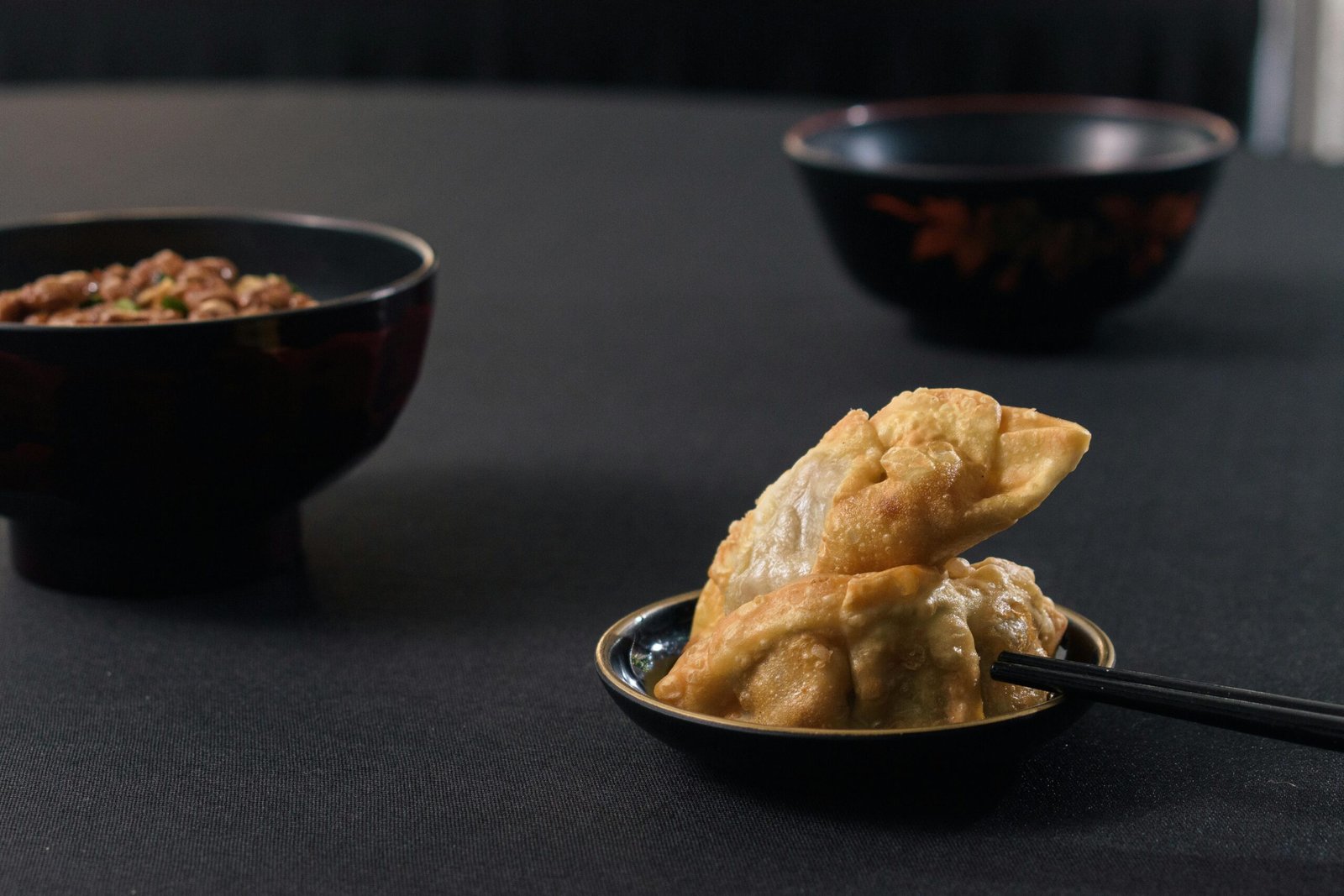 Appetizing fried dumplings with chopsticks served next to intricate black bowls on a black background.