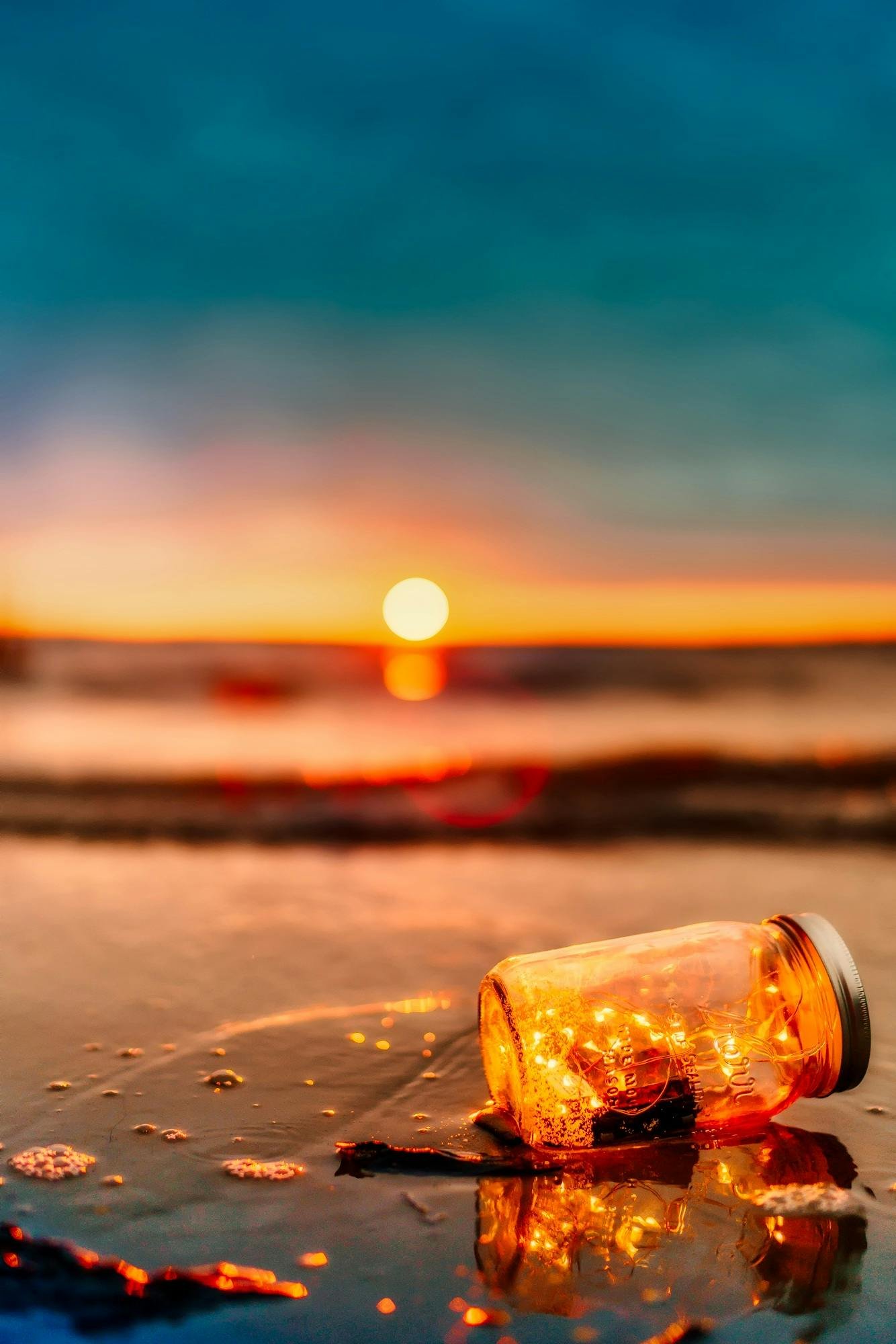 A lit jar with lights sits on a beach, creating a tranquil scene during sunset.