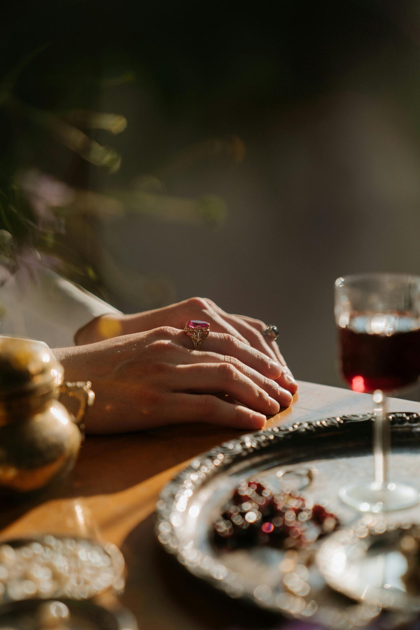 Close-up of elegant hands adorned with jewelry beside a glass of red wine on a luxurious table setting.