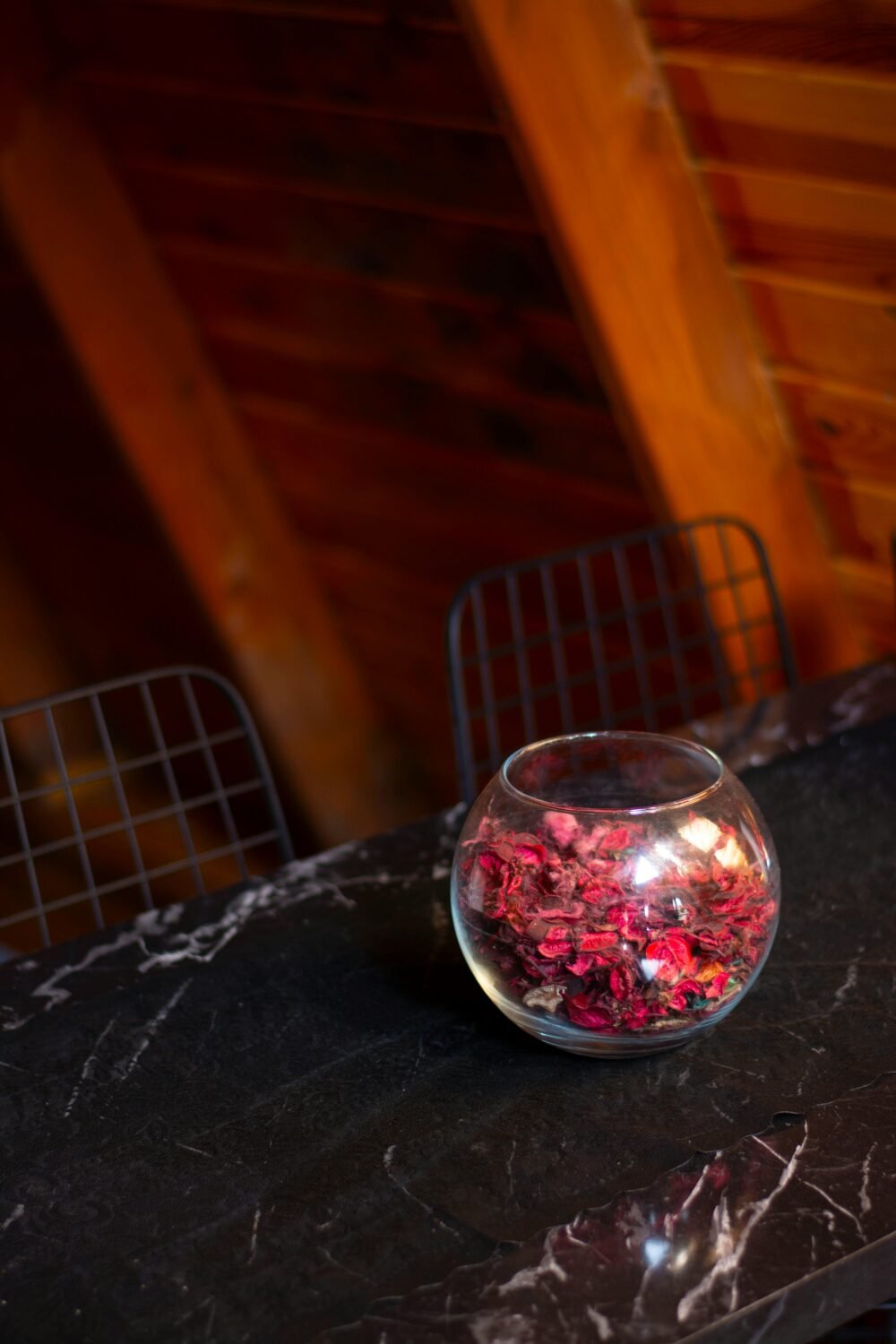 A close-up of a glass bowl filled with red petals on a black marble table in a warm wood interior.